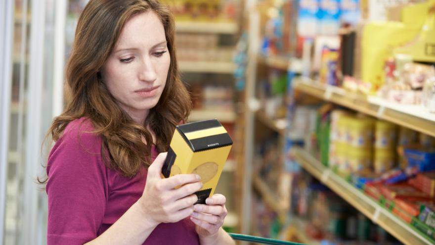 Eine Frau steht vor dem Supermarktregal, hält eine Verpackung in der Hand und schaut sie sich an.