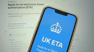 Ein Smartphone zeigt die UK-Regierungswebsite zur Beantragung der elektronischen Reisegenehmigung (ETA) für das Vereinigte Königreich an. Im Hintergrund ist ein verschwommener Bildschirm mit weiteren Informationen zur ETA sichtbar.
