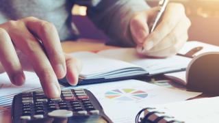 Jemand tippt auf einem Taschenrechner und schreibt dabei auf ein Blatt Papier